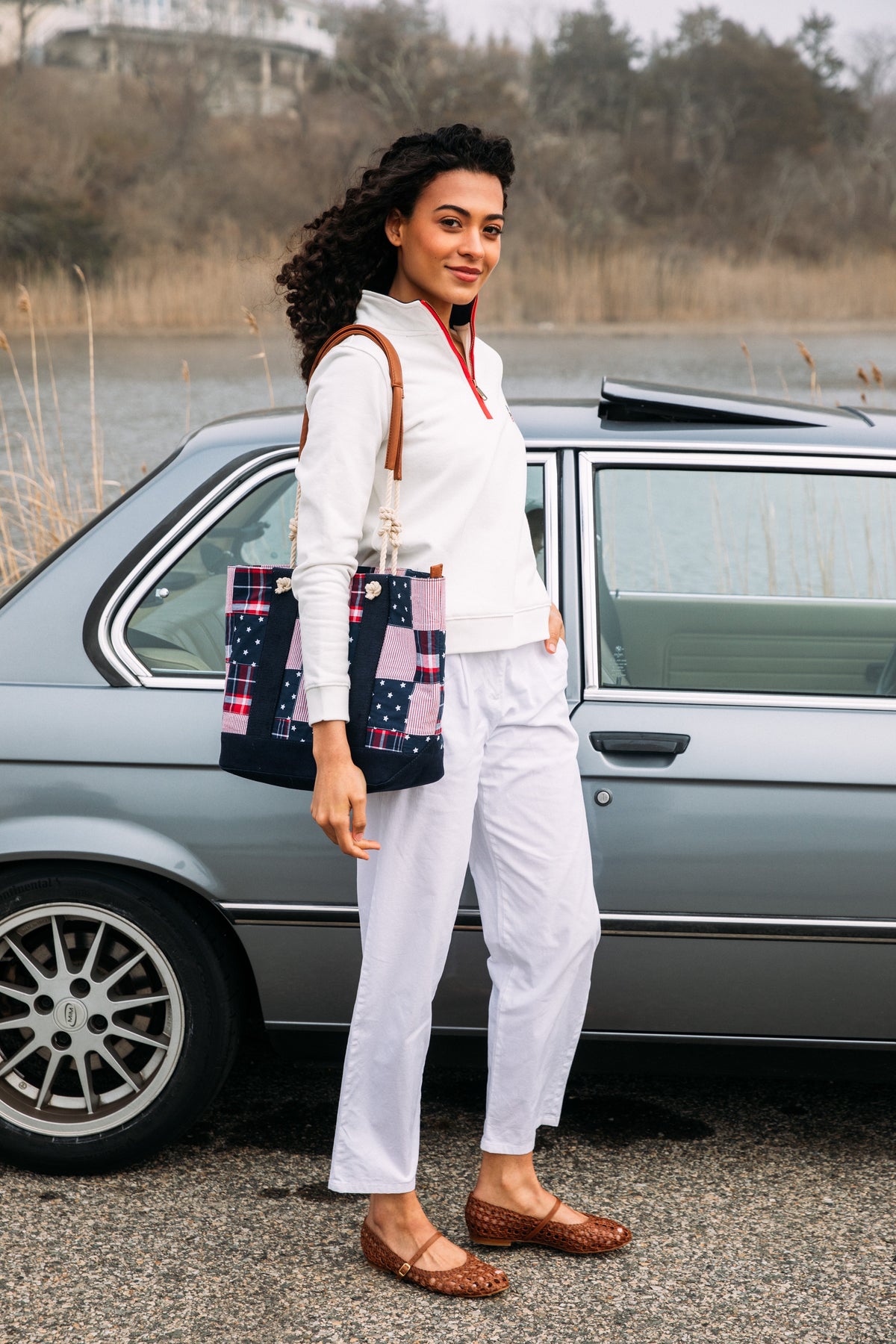 Woman in white outfit with plaid bag standing next to a vintage car by a body of water.